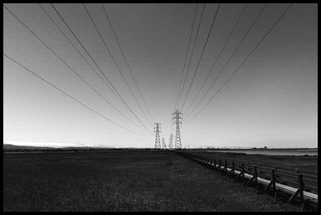 A monochrome picture of high voltage electrical wires and pilons cutting across a restored wetlands on the western shores of San Francisco Bay. The power lines and a service walkway head off into the distance across the grassy plain.

Taken May 17th 2024 at the Palo Alto Baylands Nature Preserve.