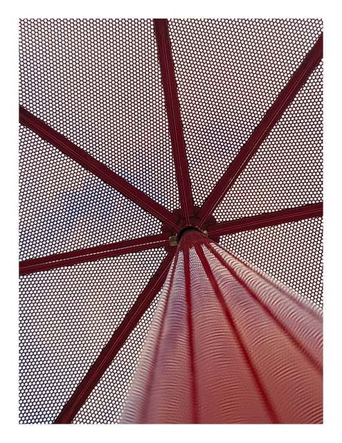 daytime. the view up a round, red, metal picnic table umbrella through the underside of its red, metal mesh, ribbed canopy to a cloudy sky with a bit of blue peeking through