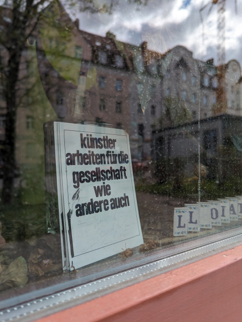Through the window of an gallery we look at a sign with black letters on white background. It reads (translated from German) "artists work for society just like others".