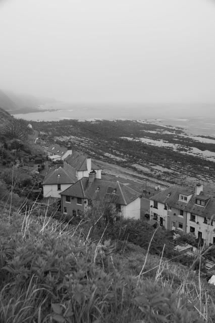 A photo taken from a cliff-side trail, looking down over a small set of 50s era houses and up towards the sea, where a bay is visible towards the horizon. There is a light fog over the sea.