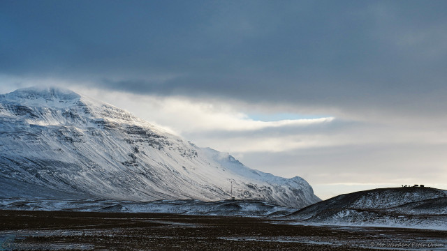 A photo of a snowy landscape. The sky is full of dense, woolly cloud but with some small windows of cyan. On the left is a mountain, its craggy flanks covered in ice and snow. In the foreground is a grey stretch of gravel or dark vegetation, and some lumpy hills. On the right, at the top of the highest hill, is a herd of horses, about 7 or 8 are silhouetted. The scene is lit from above right, and the overall impression is quite dark and gloomy.