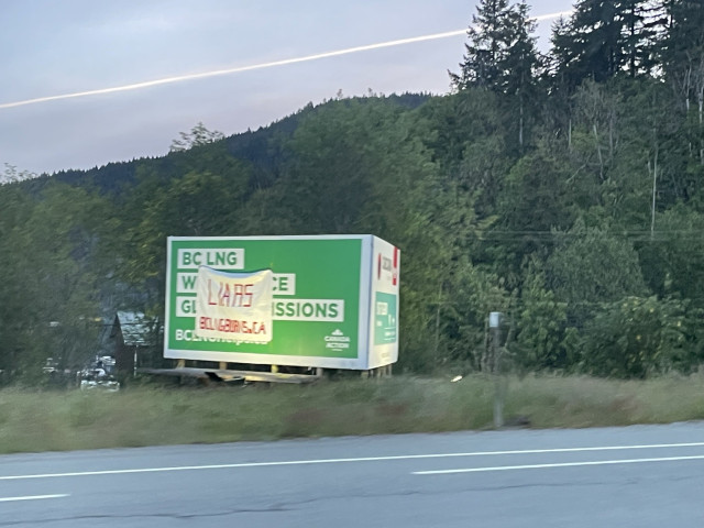 A large green billboard promoting BC LNG saying they “will reduce global emissions” has a large white tarp overtop the letters with “LIARS” in red and “BCLNGBURNS.CA”