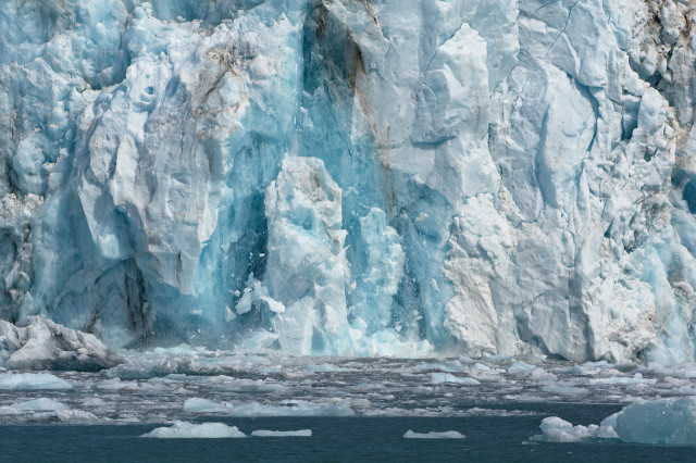 A dynamic scene of ice calving from the Columbia Glacier in the Chugach Mountains, located in the Valdez Region of Alaska during summer. The glacier's face is a jagged expanse of brilliant blue and white ice, with large chunks breaking off and crashing into the icy sea below, creating a spray of smaller ice pieces. The foreground shows a field of floating ice debris, emphasizing the glacier's active state.