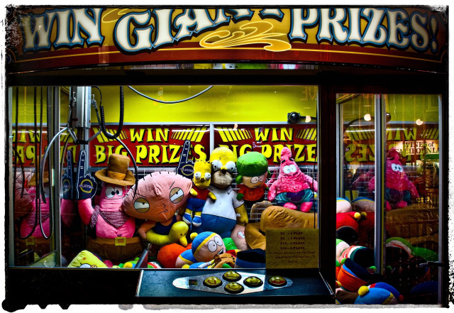 A view through the glass window of an arcade game, where you can use a claw to try and grab stuffed plush toys.

WIN GIANT PRIZES the game says. In this case the prizes are all anxious-looking characters from the Simpsons, South Park, Futurama, etc.