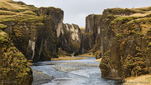 A photo of a canyon with a river running through it. Gravel islets are visible. The canyon walls are brown and grey rock, covered in mossy vegetation on the more horizontal slopes. These walls are quite vertical, in some places leaning in towards the canyon. Along the right side, at the top, seven or eight people can be seen on the grassy slopes, lending scale. The sky is uniform grey and the light is quite diffuse. It's dramatic, and beautiful.