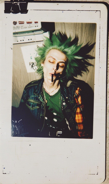 A stylized photograph of a person with green spiky hair and dramatic face makeup, evoking a punk rock aesthetic. The makeup includes black lines and shading that create a striking, almost skeletal look. The person is wearing a green shirt with a graphic design, layered with a denim jacket that has patches and a plaid shirt underneath.

A noticeable detail is the joint in their mouth, adding to the rebellious and edgy vibe of the image. The background suggests a casual setting, with some equipment and a piece of paper with text partially visible. The photograph is framed in a way that mimics an old Polaroid picture, adding a vintage feel to the image. The overall vibe of the photograph is edgy and rebellious, capturing the essence of punk culture.