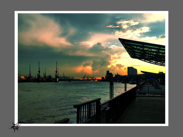 Foto aus dem Hamburger Hafen, von den Landungsbrücken geht der Blick Flussabwärts. Die Silhouette der Hafenkräne und der Häuser auf der anderen Seite schmiegen sich im spektakulären Abendrot an die Elbe. Das Abendrot und der noch blaue Himmel brechen durch durch dicke Wolken.