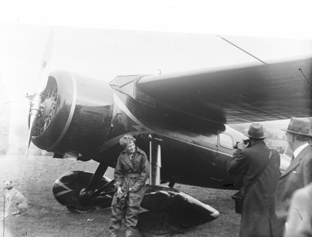 Amelia Earhart sitting on the wheel of her small prop plane, posing for photos. She's a white woman with cropped hair and dressed in a flight suit.