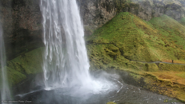 A photo taken from a high viewpoint of a landscape containing a lovely horsetail waterfall dropping into a deep plunge pool. The water is white all the way down to the point where it meets the pool, and a fan of spray and white water shows the force at which it strikes the surface. The hillsides around are green with vegetation, and there is a path leading along the side of the pool towards a cave at the back of the drop. On this path is a small figure in a black coat, demonstrating scale.
