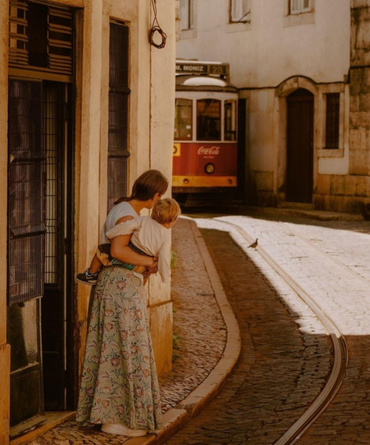 Photography. A color photo of a street scene with a mother and a small child in the narrow streets of Lisbon. The two are seen from behind. A young woman with a ponytail hairstyle, white T-shirt and flowered maxi skirt holds her baby in her arms and looks around the corner at the approaching red streetcar. She is standing in front of a store and the street is very narrow. Only a narrow sidewalk separates her from the streetcar tracks. A warm photo that conjures up different stories in the mind.