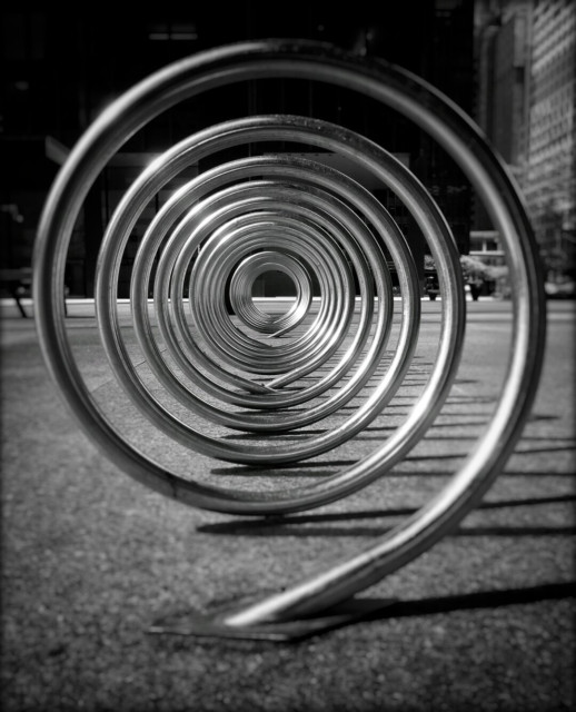 Crouched down looking through a continuous spiral bike rack. This black and white image shows the suns reflection off the shiny chrome and large buildings can be seen out of focus in the background.