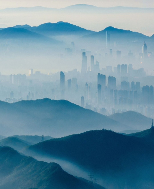 Photography. An almost surreal-looking color photo of Hong Kong. The sprawling metropolis was covered in a layer of fog, creating surreal silhouettes of the skyscrapers. Photographed from a high vantage point, the hills and skyscrapers of Hong Kong are bathed in various shades of blue. The fog and the sparse sunlight transform the view of the city into an enchanting blue watercolor painting.
Info: The image was taken from the Tai Mo Shan, the highest point in Hong Hong Kong on 23.12.2023, and is only slightly enhanced, not further processed or distorted by Photoshop.