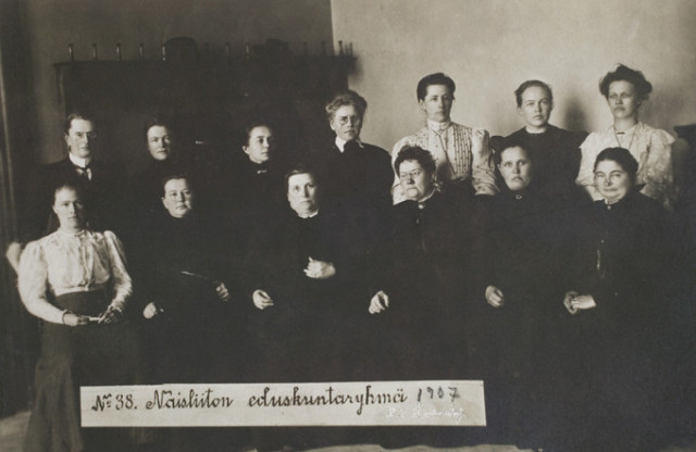 Thirteen of the nineteen women parliamentarians: six are seated and seven are standing behind them. They are all white women and most are wearing dark clothes.