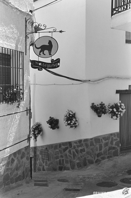A black-and-white film photo of a narrow street/alley junction with a sign which translates as "black cat corner".