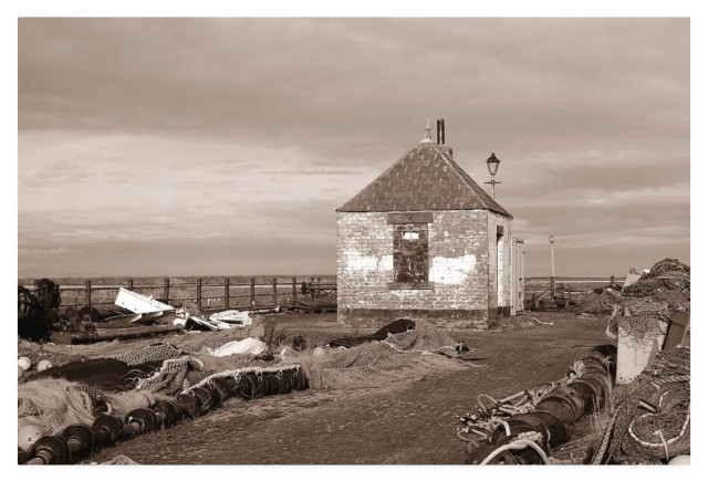 Sepia photograph showing a small stone building on a promenade surrounded by nets and floats.