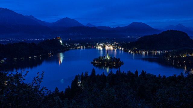 View of a lake from a hill. There is a small island in the lake. There is a church on this island. Mountains rise up behind the lake. It is blue hour.