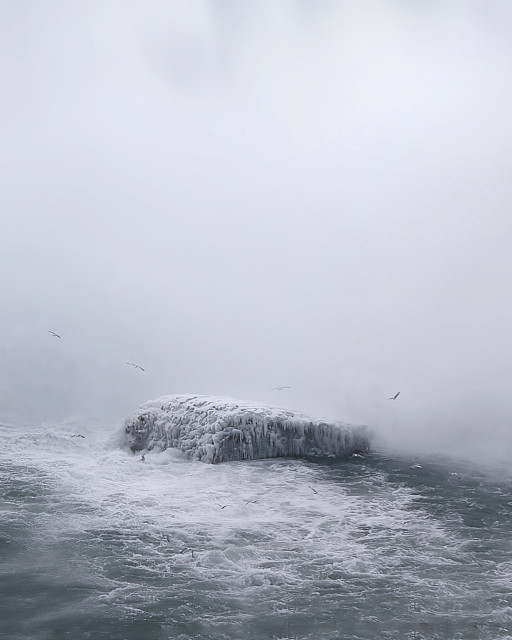 An ice covered rock stis at the bottom of a waterfall engulfed in mist. Birds fly above.