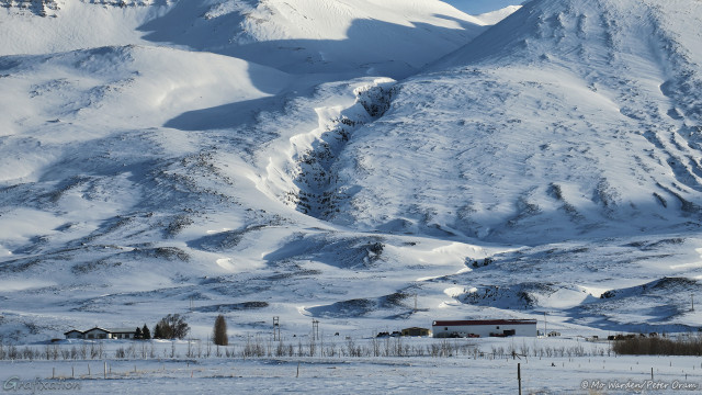 A photo of a snow scene, and the flank of a steep mountain lit from the right. A deep gulley has been created by water erosion and it curves down to ground level. At the foot are snow-covered red-roofed farm buildings, in the adjoining fields are horses. The sunlight is casting sharp shadows across the snowfields.