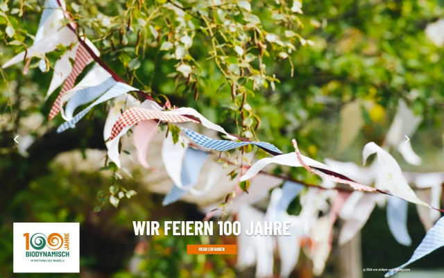Bild von der aktuellen Demeter-Webseite am 24. Mai 2024 mit dem Text "Wir feiern 100 Jahre" und dem Siegel "100 Jahre Bipdynamisch". Auf dem Foto sieht man ein Band mit bunten Wimpeln vor grünen Bäumen.