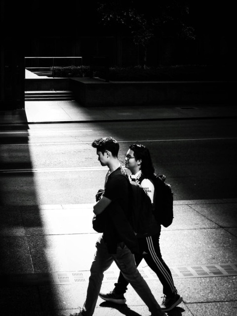 A black and white picture of two people walking along a sidewalk as a beam of light shines from between the buildings on the opposite side of the road.