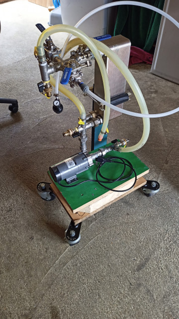 A wooden base on casters with a pump and heat exchanger bolted to it. Various pipes and valves are attached.