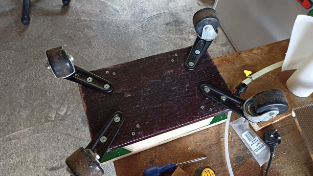 A workbench with a block of wood on it. Four caster wheels bolted to metal mounts are screwed into the wooden base.