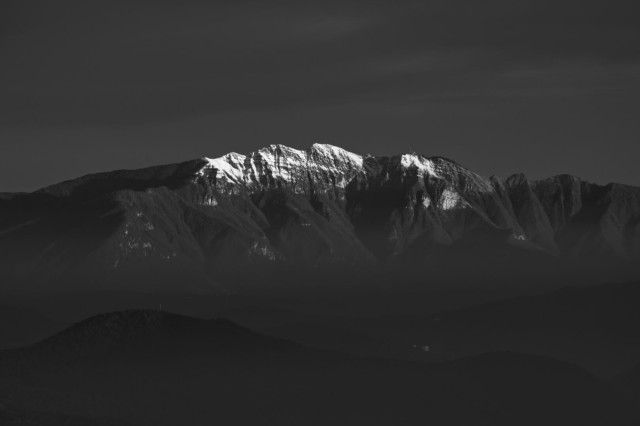 Black and white landscape photo of a mountain. The peak of the mountain is covered in snow. Smaller hills peaking out of a misty landscape fill the foreground. The sky is dark gray with only few very soft clouds.