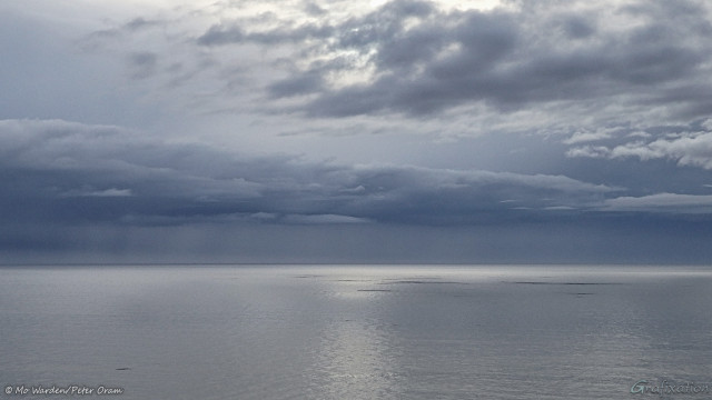A photo of a seascape. The light is from the top centre but is very diffuse. The sky looks stormy but is actually quite clear with strips of dense cloud, the background is more pearly grey than cyan. The sea is only slightly rippled, some low rocky skerries can be seen further out into the bay. The horizon is a dark line, and patches of light are visible on the water. The whole scene is in shades of silvery greyish cyan with a central stripe down the surface of the sea. It's peaceful, and very still.