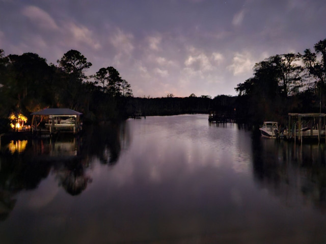 Late night overlooking a calm dark waterway where boat docks line either shoreline beneath a cloudy night sky. The left shoreline has a yellowish night light, illuminated like a campfire. The shorelines, clouds, and lighting all reflecting mirror like images on the water's surface below.
