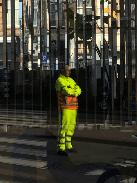 A man in a bright yellow and orange reflective road worker's outfit is seen in the middle of the frame, arms crossed and looking towards the right side of the frame. The photo is a reflection of a street scene. In the window, many vertical mirrors are visible.