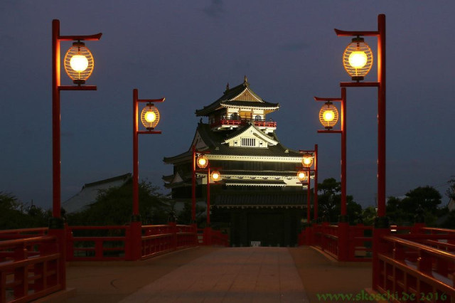 Acht rote Laternen an auf einer roten Bogenbrücke. 
Die Laternen haben die Form von Lampions .
Am Ende der Brücke befindet sich eine japanische Burg.
Es wird gerade Nacht. Die Burg ist angestrahlt. 