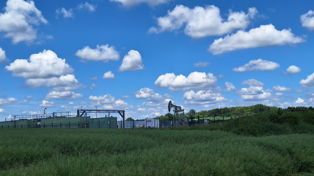 Blauer Himmel mit Wolken, grün wuchernde Wiese, eine eingezäunte Ölpumpe.