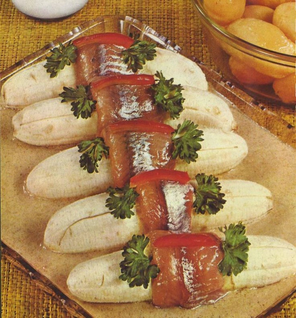 Photo from a vintage cookbook: raw bananas wrapped with slices of fish and garnished with parsley and strips of pimento.