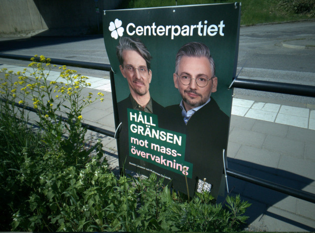 Photo of an election poster on a fence with flowers and bushes around it. The poster says  "Centerpartiet" on top, and then "HÅLL GRÄNSEN mot mass-övervakning" which means approximately "stand firm against mass surveillance". The poster shows the faces of two persons and at the bottom of the poster are two names, Svante Linusson and Muharrem Demirok