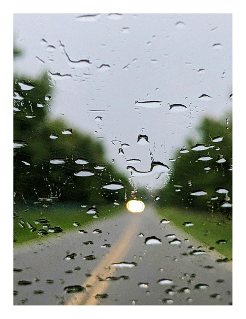 abstract. stormy afternoon. view through raindrops on my windshield. out-of-focus, two-lane road with grass and trees along both sides. in the mid-distance, a car approaches in the opposite lane with headlights on.