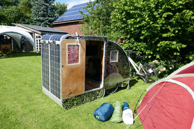 A bicycle camper on a meadow with tents. The trailer expands by folding it out. The outer shell folds over a smaller inner section or is folded down or up for use. This doubles the length and an adult can fit inside comfortably