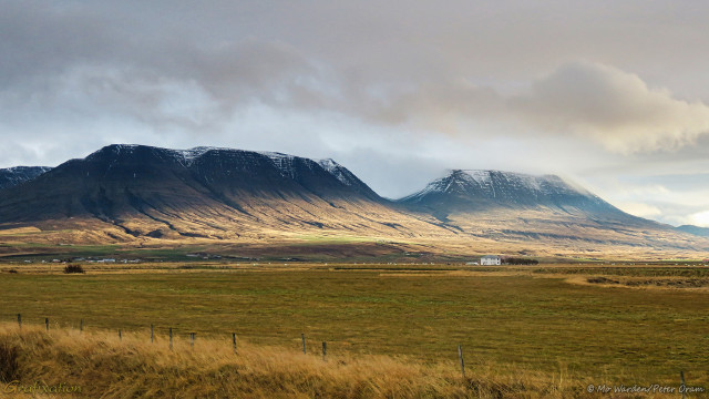 The same two mountains, taken from slightly nearer than the first shot. This time the ground is free from snow and is covered in golden vegetation, suggesting autumn. The lower slopes of the two mountains are touched by sunlight from the right, and clouds have created shadow across the flat tops of the peaks. The bare rock is dark grey, and a sprinkle of snow is visible on the tops. The sky is cloudy again but with some clearer patches.