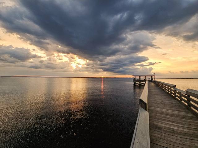 Wide view over a vast, calm river beneath huge grey cloud formations with the setting sun behind, casting a red glow across the water's surface and bright yellow and orange rays between cloud openings, reflecting upon the water below. With a view on the right of the very long, weathered, wooden fishing pier and covered observation deck reaching far out into the river.
