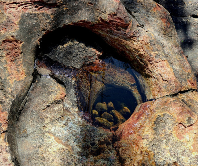 Photograph of a giant's kettle in the grey, orange and yellow rock surface of a riverbank. The round shape of the kettle is clearly seen, with small rocks in it and remaining water reflecting a little bit of blue sky. The whole area was flooded a couple of weeks ago.