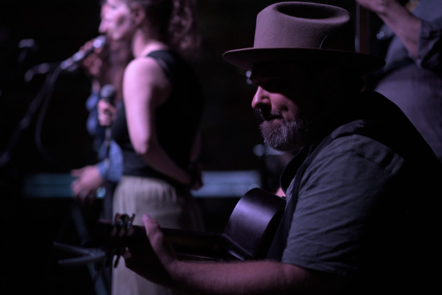 Ein Mann mit Akustikgitarre sitzt auf einer Bühne. Sein Gesicht ist halb abgedunkelt weil er einen großen Hut trägt

Im Hintergrund sind weitete Musiker innen zu erkennen