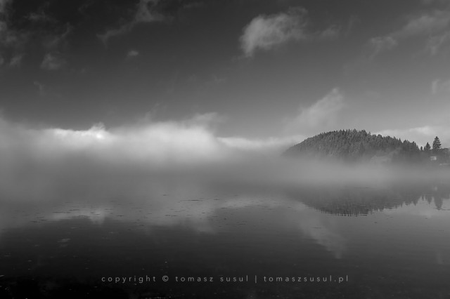 A black and white photograph of a lake with a thick, low mist flowing from the left, illuminated from above by the rising sun. On the right is a patch of land with a hill still rising above the fog. The calm surface of the lake creates a mirror in which the mist and small clouds in the sky are reflected. The fog obscures the boundary between water and land.