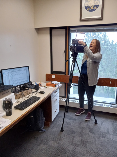 A view of my office with a journalist laughing as she sets up a camera on a tripod in front of the office window, with a nice view of trees.  There's a big monitor and some clutter on my big white desk.