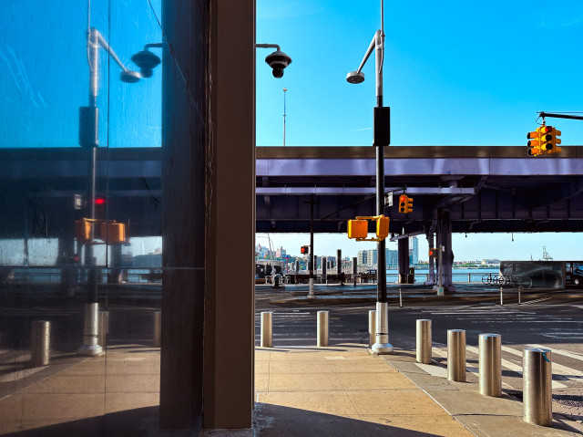 Street scene with traffic lights, security cameras, and a view of a waterfront. A reflective stone wall splits the image, capturing the urban infrastructure under a clear blue sky.