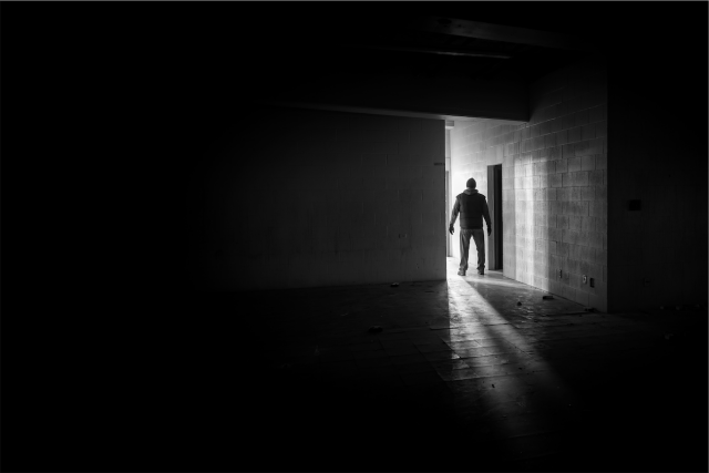 The image is a black-and-white photograph depicting a person standing in a dimly lit hallway. The person is silhouetted against a bright doorway, creating a stark contrast between light and shadow. The individual appears to be wearing a hoodie and pants, with their hands slightly away from their sides. The hallway is empty, with light streaming in from the doorway, casting long shadows on the floor. The walls are made of large, rectangular tiles, adding to the stark, austere atmosphere of the scene. The overall mood of the image is dark and mysterious, enhanced by the high contrast and the person's ambiguous posture.