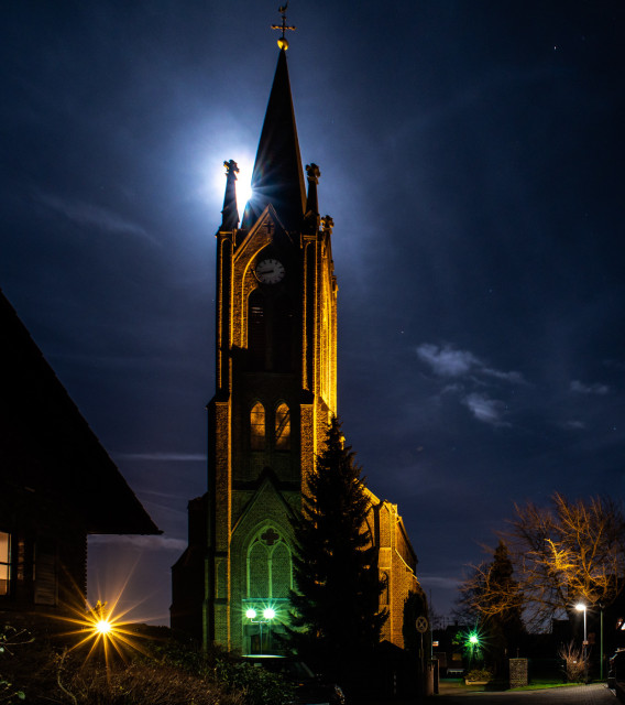 Das Nachtfoto zeigt die Kirche St.Pankratius in Glessen, dem Dorf in dem ich wohne. Der Mond bestrahlt den Himmel und zeigt sich hinter der Spitze des Turms. 
Sie wird nachts gelb/orange angestrahlt, unten ist ein grünes Licht zu sehen. Gebäude und ein paar Bäume sind rechts und links unten zu sehen. 