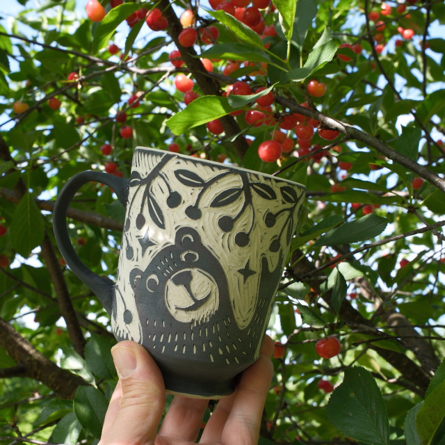 a photo of an unfired cup in front of a cherry tree, filled with fruit. The cup is decorated with a black bear picking cherries