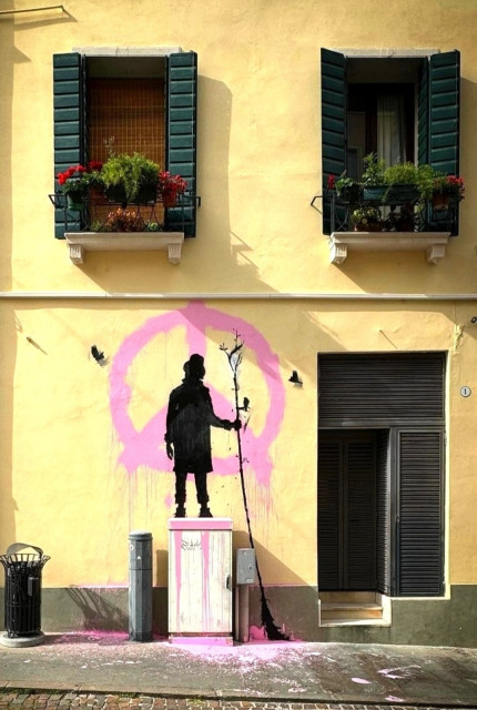 Streetartwall.  A mural with the silhouette of a man and a peace sign was sprayed onto a yellowish house wall. It is the black silhouette of a man in a coat and hat standing over a (real) gray distribution box. He is holding a long tree trunk with roots in his hand and looking at his work. Three painted birds are flying around. He has painted a pink "peace sign" on the wall and the paint is dripping onto the floor. Simple and magnificent.