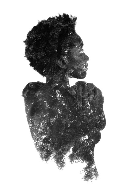 A dark skinned woman is standing naked with her back to the camera. Her lower back fades into leaves and trees and her skin and face are specked with points of light, like rays peeking through the leaves. Black and white. 