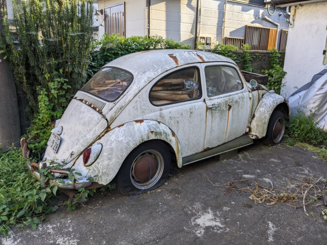 Alter, rostiger, weißer VW Käfer von schräg hinten.
Die Reifen sind platt.
Das Innere mit Holzbalken gefüllt.

