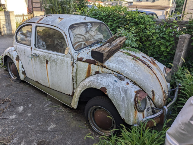 Alter, rostiger, weißer VW Käfer von schräg vorne.
Die Reifen sind platt.
Das Innere mit Holzbalken gefüllt.